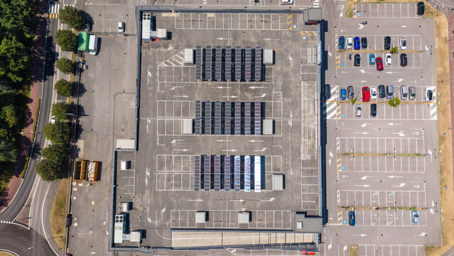 Aerial view of a shopping mall rooftop featuring rows of solar panels, adjacent parking areas with marked spaces, and surrounding greenery in Peschiera Borromeo, Lombardy, Italy.