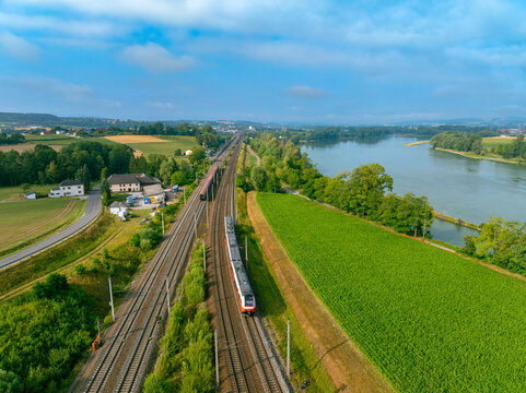 Aerial view of local trains traveling along the Danube river through green fields and rural landscape in Sarling, Lower Austria, Austria.