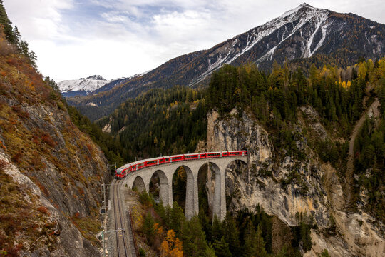 View of the iconic red train crossing the Landwasser Viaduct, surrounded by steep rocky cliffs, dense pine forests, and snow-capped mountains in Switzerland.