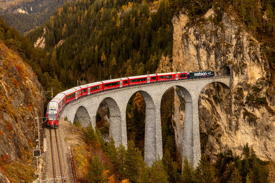 View of the Landwasser Viaduct with a red train crossing the stone arch bridge surrounded by autumn forest and rocky cliffs in Switzerland.