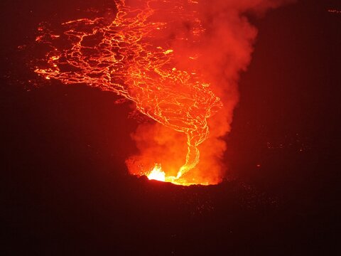 Aerial view of a volcanic eruption with glowing lava flowing from the crater, creating a bright orange contrast against the dark volcanic landscape in Iceland.
