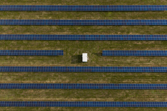 Aerial view of rows of solar panels arranged in a grassy field with a small white utility building in the center in Poland.