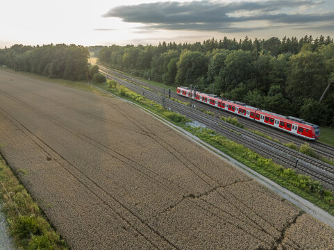 Aerial view of a red and white commuter train traveling on tracks alongside a golden wheat field and a dense forest in Germany.
