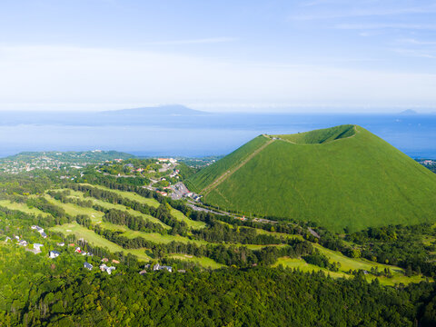 Aerial view of Mt. Omuro, a prominent green volcanic cinder cone, surrounded by lush forests, a golf course, and the blue ocean in the background in Ito, Shizuoka, Japan.