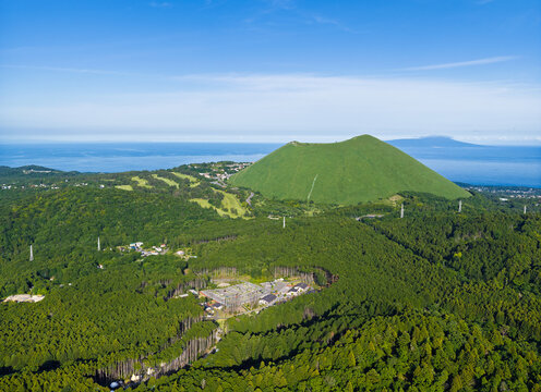 Aerial view of Mt. Omuro, a dormant volcanic cone covered in lush green grass, surrounded by dense forests and the blue waters of the Pacific Ocean, Ito, Shizuoka, Japan.