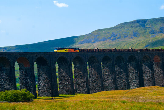 View of the Ribblehead Viaduct with a freight train crossing the stone arches against a backdrop of green hills and a clear blue sky in Carnforth, England, United Kingdom.