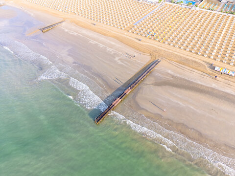 Aerial view of the sandy beach and wooden pier extending into the turquoise sea, with rows of sun umbrellas on the shore in Lido di Jesolo, Veneto, Italy.