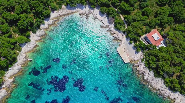 Aerial view of Pakleni Islands coastline with turquoise water, a small stone pier, and a house with a red roof nestled in pine trees, Split-Dalmatia County, Croatia.