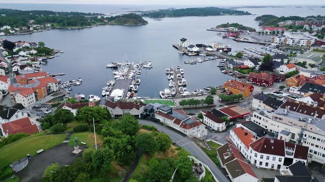 Aerial view of the coastal town of Grimstad featuring a busy harbor filled with boats, colorful houses, and green hills in Grimstad, Agder, Norway.