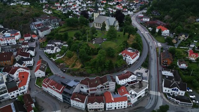 Aerial view of the historic Grimstad church surrounded by lush greenery, residential rooftops, and winding roads in the coastal town of Grimstad, Agder, Norway.