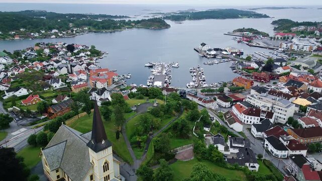 Aerial view of Grimstad Church overlooking the harbor with boats, colorful rooftops, and lush greenery in Grimstad, Agder, Norway.