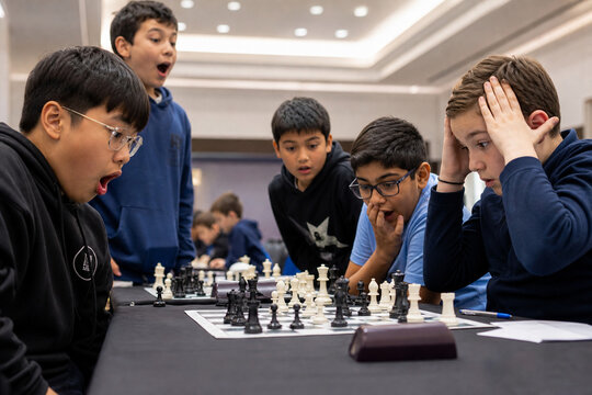 A group of young students watches an intense chess match at a tournament, surprised and focused. The bright classroom setting highlights strategy, concentration, and learning through game play.