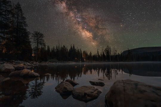 View of the Milky Way core shining brightly above the pine trees and reflecting on the calm surface of Mirror Lake in Duchesne County, Utah, United States.