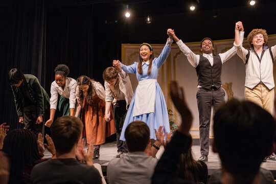 Diverse actors bow on stage after a successful theatrical performance. The happy cast holds hands and smiles at the audience while receiving applause. Culture and art concept.