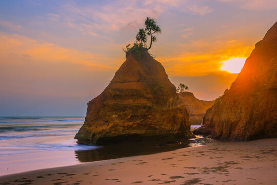 View of a unique coastal rock formation with trees on top at sunset, featuring a sandy beach and gentle waves in Lais, Bengkulu, Indonesia.