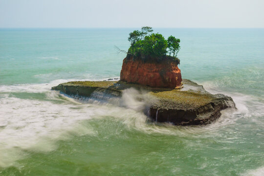 View of a small rocky island with lush green trees and reddish cliffs surrounded by crashing waves in the ocean in Lais, Bengkulu, Indonesia.