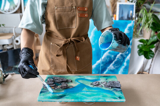 Woman creative artist working on handmade liquid art painting in home workshop at worktable, craftswoman holding cup with resin and pouring liquid on canvas, using tools to evenly distribute resin.