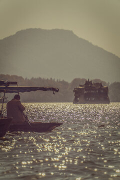 View of a boatman rowing a traditional wooden boat on the shimmering waters of West Lake, with a pavilion boat in the distance against a hazy mountain backdrop in Hangzhou, China.