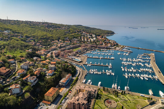 Aerial view of Muggia harbor with sailboats docked in the marina, surrounded by coastal buildings, lush green hills, and the blue Adriatic Sea in Trieste, Friuli-Venezia Giulia, Italy.