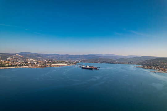 Aerial view of the Gulf of Trieste featuring the town of Muggia, industrial port infrastructure, and calm blue waters under a clear sky in Trieste, Friuli-Venezia Giulia, Italy.