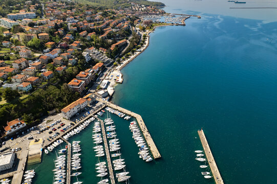 Aerial view of Muggia with its marina filled with sailboats, historic buildings with terracotta rooftops, and the blue waters of the Adriatic Sea in Trieste, Friuli-Venezia Giulia, Italy.