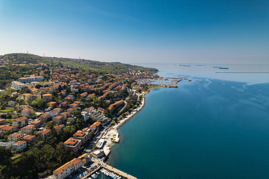 Aerial view of Muggia harbor with boats docked along the coastline, residential buildings, and green hills overlooking the Adriatic Sea in Trieste, Friuli-Venezia Giulia, Italy.