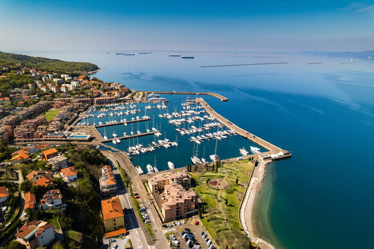 Aerial view of Muggia marina with sailboats and yachts docked in the harbor, surrounded by coastal town buildings and the blue sea in Trieste, Friuli-Venezia Giulia, Italy.