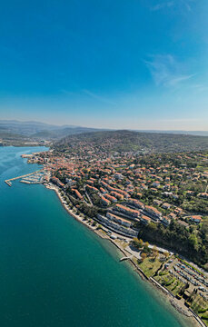 Aerial view of Muggia, a coastal town with terracotta-roofed houses, a marina, and green hills overlooking the sea in Trieste, Friuli-Venezia Giulia, Italy.