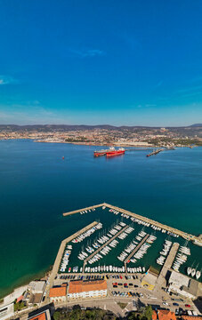 Aerial view of Muggia marina with sailboats and motorboats docked in clear blue water, surrounded by coastal buildings and a distant shoreline in Trieste, Friuli-Venezia Giulia, Italy.