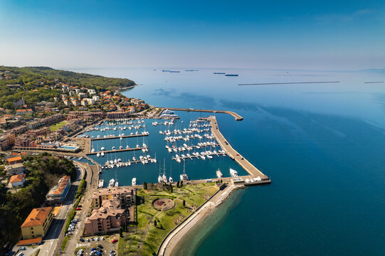 Aerial view of Muggia marina with sailboats and yachts docked in the harbor, surrounded by coastal buildings and hills along the Adriatic Sea in Trieste, Friuli-Venezia Giulia, Italy.