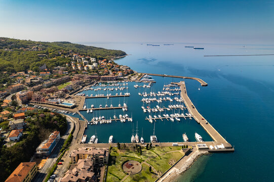 Aerial view of the Muggia marina filled with numerous yachts and sailboats, situated along the coastal town with historic buildings and lush greenery, Trieste, Friuli-Venezia Giulia, Italy.