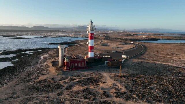 Aerial view of the iconic red and white striped Faro de Toston lighthouse standing on the rugged rocky coastline during golden hour in La Oliva, Canary Islands, Spain.