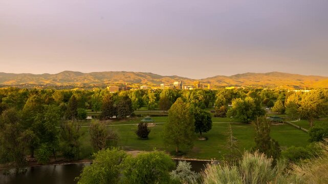 Aerial view of Ann Morrison Park featuring lush green trees, a river, and the city skyline under golden hour light in Boise, Idaho, United States.