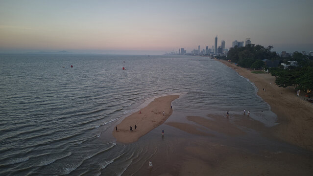 Aerial view of a sandbar at low tide with people walking along the shore and a distant city skyline under a soft evening sky in Ao Nang, Krabi, Thailand.