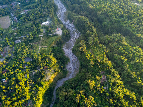 Aerial view of the Turgo river valley featuring a rocky riverbed winding through dense tropical forest and scattered buildings in Pakem, Special Region of Yogyakarta, Indonesia.