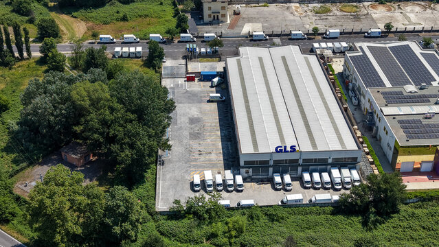 Milan, Italy - 27 May 2026: Aerial view of the GLS logistics facility featuring a large warehouse, numerous delivery vans parked in the loading area, and surrounding greenery.