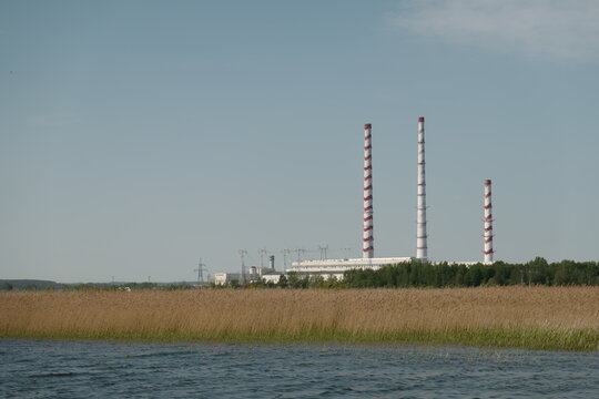 View of the Elektrenai Power Plant with its distinctive red and white striped chimneys rising above the surrounding trees and reeds near the water in Elektrenai, Vilnius County, Lithuania.