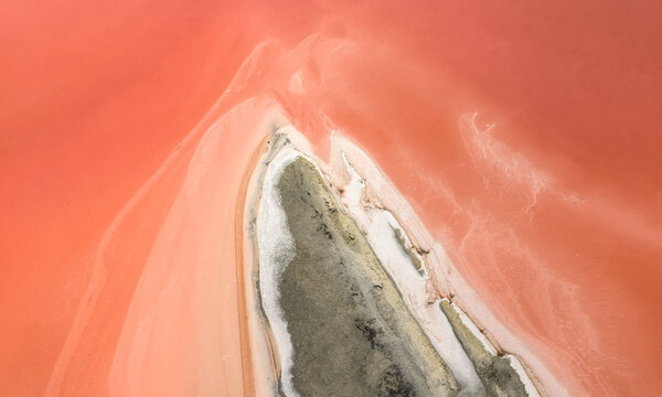 Aerial view of coral pink saltwater surrounding a mineral island with delicate white salt textures and organic flowing patterns in France.