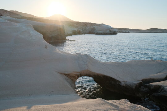 Soft sunrise light illuminates the smooth white volcanic rock and natural arch at Sarakiniko, with calm blue water and a hazy horizon creating a serene coastal scene in Milos, Greece.