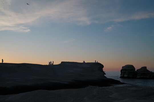 Silhouetted figures stand along smooth volcanic rock as warm sunrise light glows beneath a pastel sky, creating a quiet and atmospheric coastal scene in Sarakiniko, Milos, Greece.
