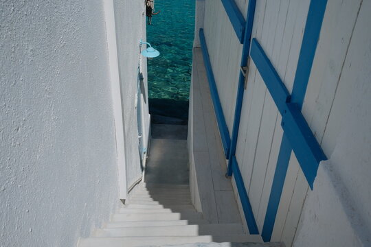 A narrow passage between whitewashed Greek buildings reveals vivid turquoise water beyond, combining blue accents, traditional island architecture and the bright colors of the Aegean.