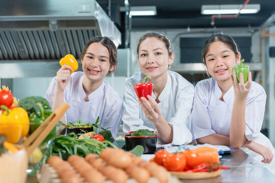 Female chef teacher and young student girls holding fresh bell peppers in cooking class commercial kitchen, Happy cooking instructor and kids showing colorful vegetables in culinary school kitchen