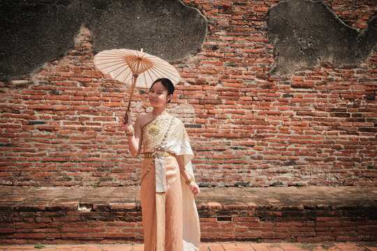 Portrait of Asian teenage girl dressed in traditional Thai outfit and umbrella with cheerful smile at old brick wall, summer holiday travel in Ayutthaya, ancient ruins heritage,  tourist destination.