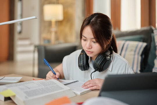 Asian teenage girl in headphones concentrates on learning at home, reading textbooks, note writing, self-tutorial exams, preparation for back to school, Homeschooling, university education lifestyle.