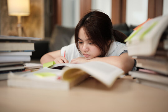 Asian teenage girl concentrates on pile of books, focuses on learning at home with pile of textbooks, reading and writing, self-study for school, Homeschooling, university education lifestyle.