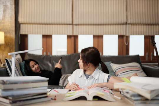 Two Asian teenage tutorials, girl concentrates on online class, remote learning at home with tablet, her friend uses mobile phone on sofa, self-study with textbooks, homeschooling together lifestyle.
