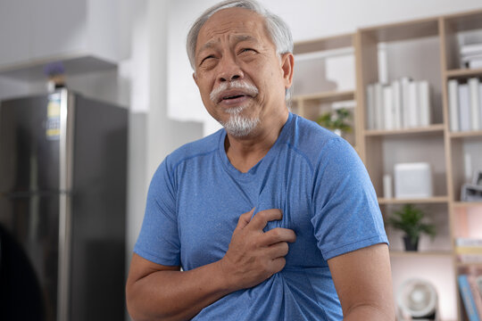 Senior asian man clutching his chest with severe pain during sudden heart attack inside home showing deep distress and fear of medical emergency indoor while breathing heavily