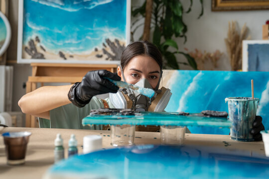 Professional craftswoman creating liquid art painting, carefully applies epoxy resin with spatula, focused female artist in safety equipment adding fluid resin on surface and controls process.