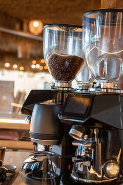 Modern black espresso coffee grinders displaying roasted beans in transparent hoppers on a cafe counter. Warm illuminated background highlighting professional barista equipment in a coffee shop.
