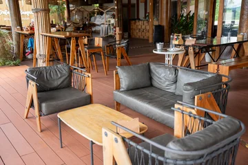 Fotobehang Horeca Empty cozy tropical cafe interior featuring a white table, woven rattan chairs with cushions, wooden flooring, and large wooden pillars against a white wall near glass folding doors.  © Yellow Boat